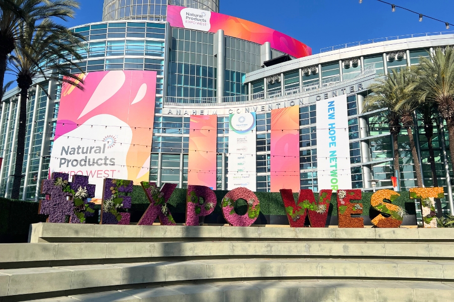 Anaheim Convention Center with signage for Natural Products Expo West and New Hope Network in the background, foreground shows an #EXPOWEST garden signage filled with succulents and flowers in the letters above tiered concrete seats.