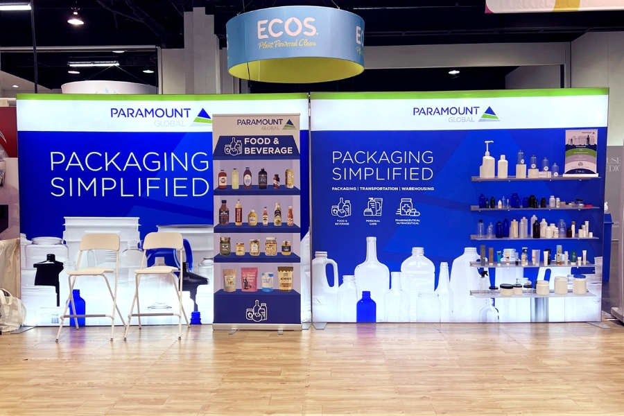 Paramount Global Booth, 2 white chairs on the left, shelves with product packaging on the right. Signage on the back drop says: Packaging Simplified with the Paramount Global logo at the top. There is a pop-up sign in the middle that show images of food and beverage packaging.